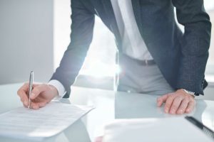 Young elegant businessman in formalwear signing financial documents while bending over desk in office